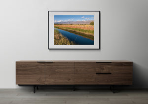 Warm grasses border a calm blue river below scattered white clouds.