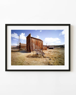 Weathered wooden buildings stand in a California ghost town beneath a blue sky and white clouds.