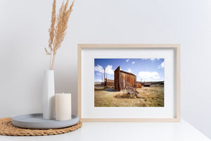 Colour photograph of rustic timber structures, open ground and a bright western sky.