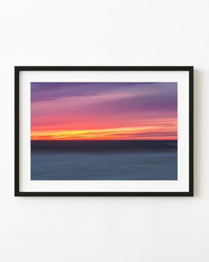 Pink and violet sunset clouds stretch above a dark California ocean and glowing yellow horizon.