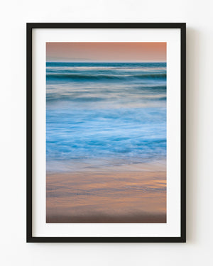 Blue Pacific waves wash over peach-coloured sand beneath a soft California sunset sky.