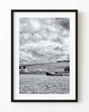Black and white Cape Breton coastal photograph with a boat, hillside home and textured clouds.