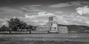 A weathered wooden grain elevator rises above railway cars beneath a cloudy prairie sky.