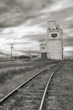 Curving rails cross open prairie toward a tall wooden elevator beneath textured clouds.