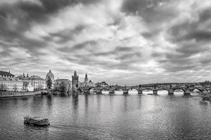 Stone bridge arches cross the river below historic Prague towers and a textured sky.