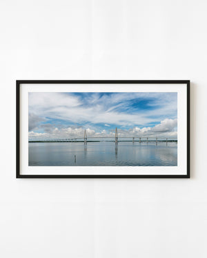 Ravenel Bridge spans calm water in Charleston beneath sweeping white clouds and a blue sky.