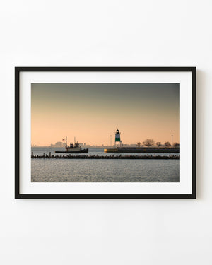 A tugboat crosses Chicago harbor beside a striped lighthouse beneath a warm evening sky.