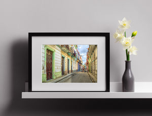Pastel Cuban buildings frame a receding street with children and a strip of cloudy blue sky.
