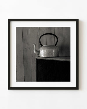 Black and white still life of a metal teapot on a dark cabinet against wooden boards in Clarence Town.