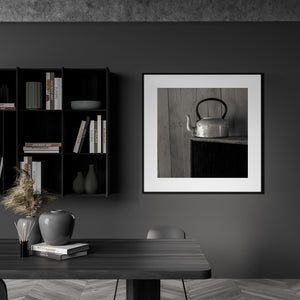 Square still life from Clarence Town showing a weathered metal kettle against a rustic wooden wall.