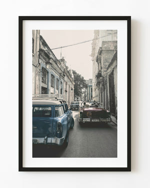 Classic cars travel through a narrow Old Havana street lined with weathered buildings in Cuba.