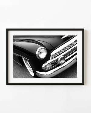 Black and white close-up of a classic Chevrolet’s round headlight, curved front fender and chrome grille.