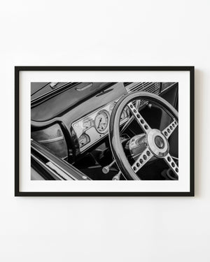 Black and white photograph of a classic Ford convertible interior with a perforated steering wheel and dashboard gauges.