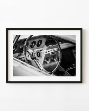 Black and white photograph of a classic Ford Mustang interior with a large steering wheel and circular dashboard gauges.