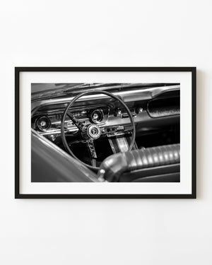 Black and white photograph of a classic Ford Mustang cabin with its steering wheel, dashboard and ribbed seats.
