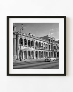 Black and white Havana street photograph with a classic car passing a long building of repeated arches and columns.