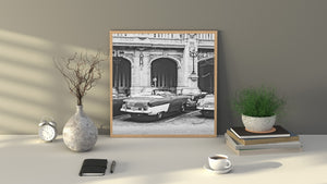 Monochrome Cuban city scene with vintage convertibles parked beside decorative lamps and arched doorways.
