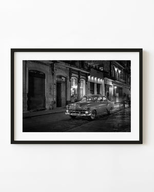 Black and white night photograph of a classic Havana taxi beside weathered buildings and illuminated doorways.