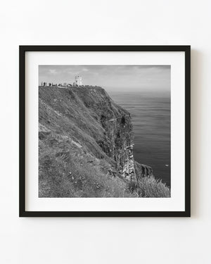 Black and white photograph of a tower above the Cliffs of Moher with grassy slopes and the Atlantic beyond.