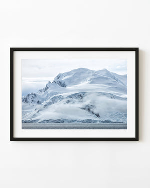 A snow-covered Antarctic mountain rises above coastal water with low clouds wrapped around its slopes.