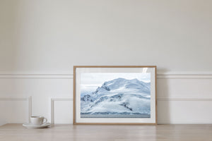 Layered snow and exposed rock form a coastal mountain beneath a pale overcast sky.