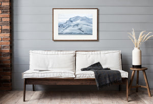 A rugged polar shoreline lies below smooth snow ridges and mist-covered mountain contours.