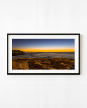 Golden dawn light stretches across Clovelly Beach above textured rocks and shallow coastal pools.