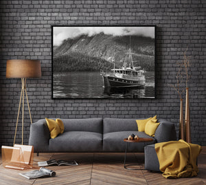 Monochrome maritime landscape with a small boat reflected on calm water beneath dense shoreline forest.