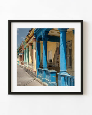 Blue columns and yellow walls line a sunlit Cuban street in this colour photograph.