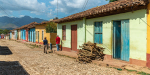 A long row of pastel Cuban houses with blue doors borders a sunlit cobblestone street.
