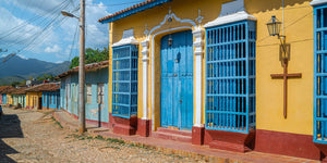 A sunlit yellow Cuban facade with tall blue windows borders a cobbled street.
