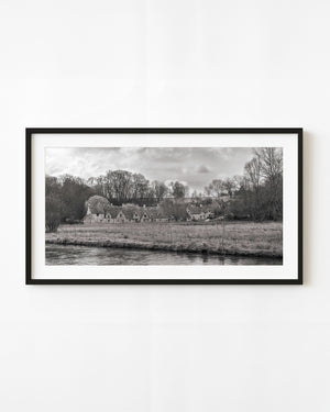 Black and white panorama of stone cottages beside a river in the Cotswolds, England.