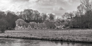 A quiet river runs before grassy banks, village cottages and trees in the English countryside.