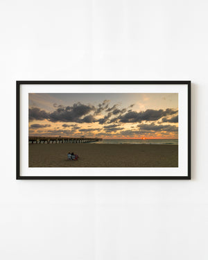 A couple sits on Juno Beach beside the pier beneath clouds lit with golden coastal light.