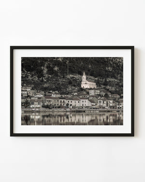 A Croatian riverside village and hillside church reflect in calm water in black and white.