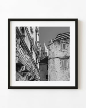 Croatian stone buildings frame a distant bell tower in a square black and white photograph.