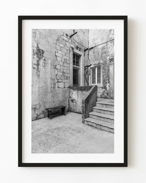 Stone steps climb beside a weathered wall and bench in a Croatian courtyard, photographed in black and white.