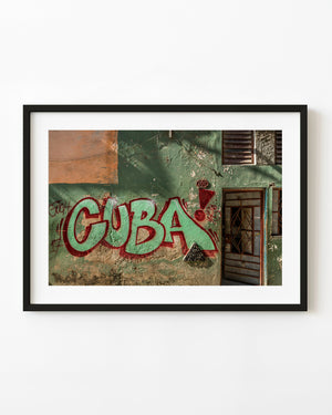 CUBA graffiti stretches across a weathered green house wall beside a barred doorway.