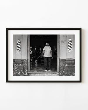 A man enters a Cuban barber shop between painted barber poles in a black and white street photograph.