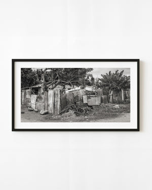 A Cuban house stands behind a patchwork corrugated metal fence in a wide black and white photograph.