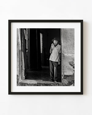 A Cuban man stands in a weathered doorway with one hand raised near his face in black and white.