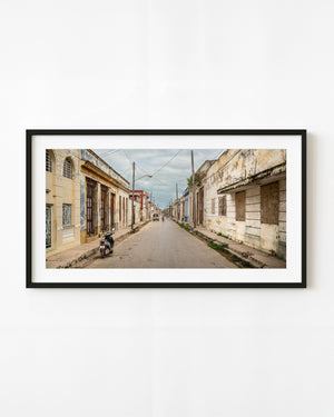 A quiet Cuban street recedes between weathered facades with a distant cyclist in a colour photograph.