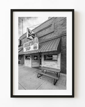 Curley’s Tap pub frontage displays a Route 66 sign above a sidewalk bench in black and white.