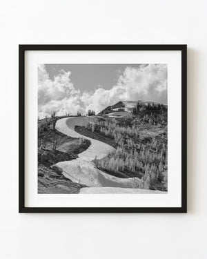 A curving snowfield winds through mountain trees beneath bright clouds in a black and white landscape.