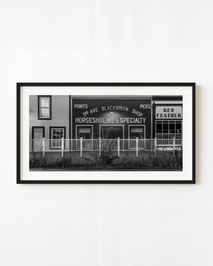 A historic Dawson City blacksmith shop displays painted trade signs behind a wooden porch in black and white.