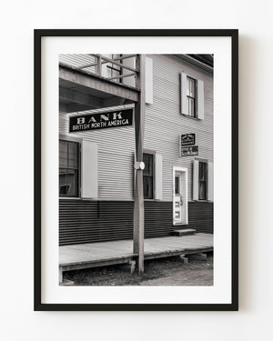 A hanging bank sign and wooden balcony frame a historic Dawson City facade in monochrome.