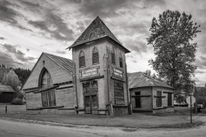 A weathered church tower and adjoining rooflines rise beside a tall tree in Dawson City.