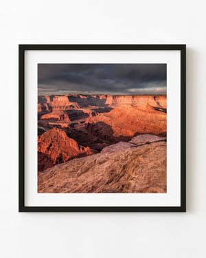 Warm orange light illuminates layered canyon cliffs beneath dark clouds at Dead Horse Point, Utah.