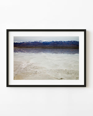 Snowy mountains reflect across shallow water beyond pale patterned salt flats in Death Valley.