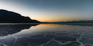 Angular pale salt patterns border shallow reflective water as evening falls in Death Valley.
